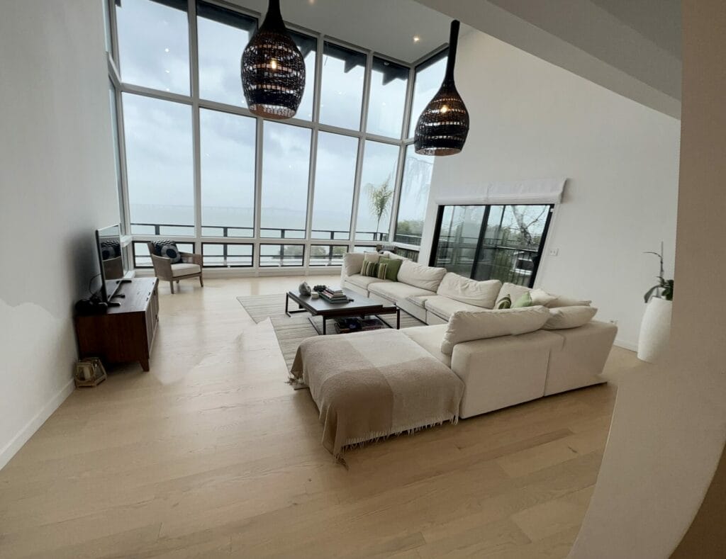 A Modern Living Room Features Large Floor-To-Ceiling Windows, A White Sectional Sofa, A Wooden Tv Stand, And A Minimalist Decor. The Space Is Well-Lit With Natural Light.