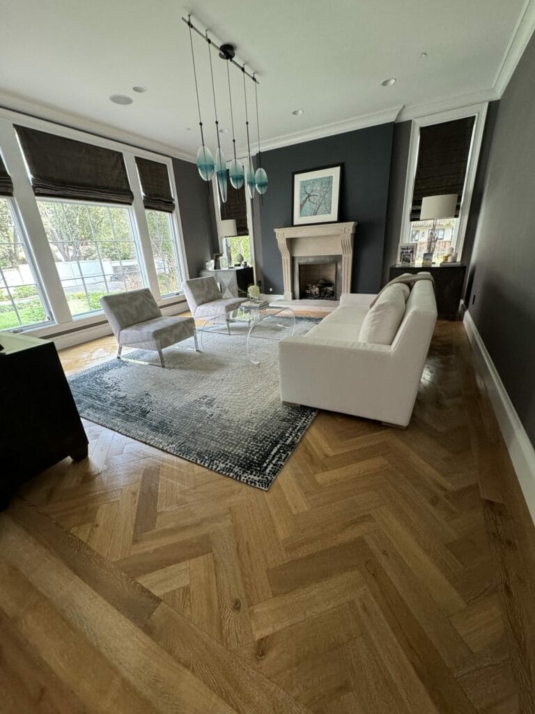 A Modern Living Room With Wooden Herringbone Flooring, A White Sofa, Two Grey Chairs, A Glass Coffee Table, A Fireplace With Artwork Above, And Large Windows With Black Shades.
